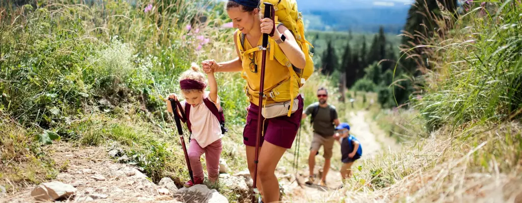 Das Bild zeigt eine Frau mit Wanderstock und Kind an der Hand. Sie laufen einen Berg hinauf. Im Hintergrund sieht man einen Mann mit Sohn, die das Ganze beobachten. Das Bild wird im Bereich "Impfschutz, der mitreist" verwendet.