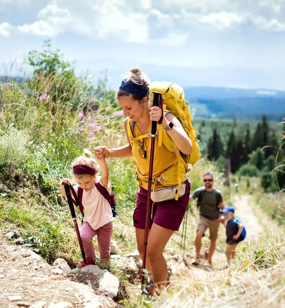 Das Bild zeigt eine Frau mit Wanderstock und Kind an der Hand. Sie laufen einen Berg hinauf. Im Hintergrund sieht man einen Mann mit Sohn, die das Ganze beobachten. Das Bild wird im Bereich "Impfschutz, der mitreist" verwendet.