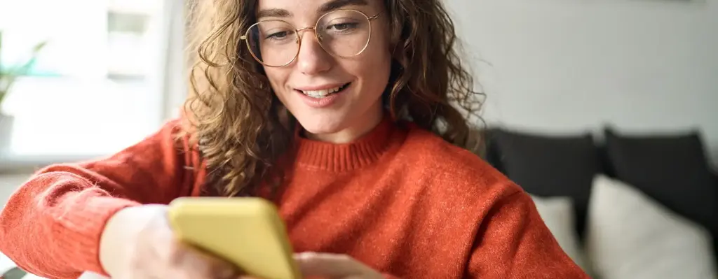 Das Bild zeigt eine junge Frau in einem rotorangen Pullover. Sie trägt eine Brille und blick auf ihr Smartphone. Das Bild wird im Bereich "Krankenkasse? Kannst du jetzt auch per Service-App" verwendet.
