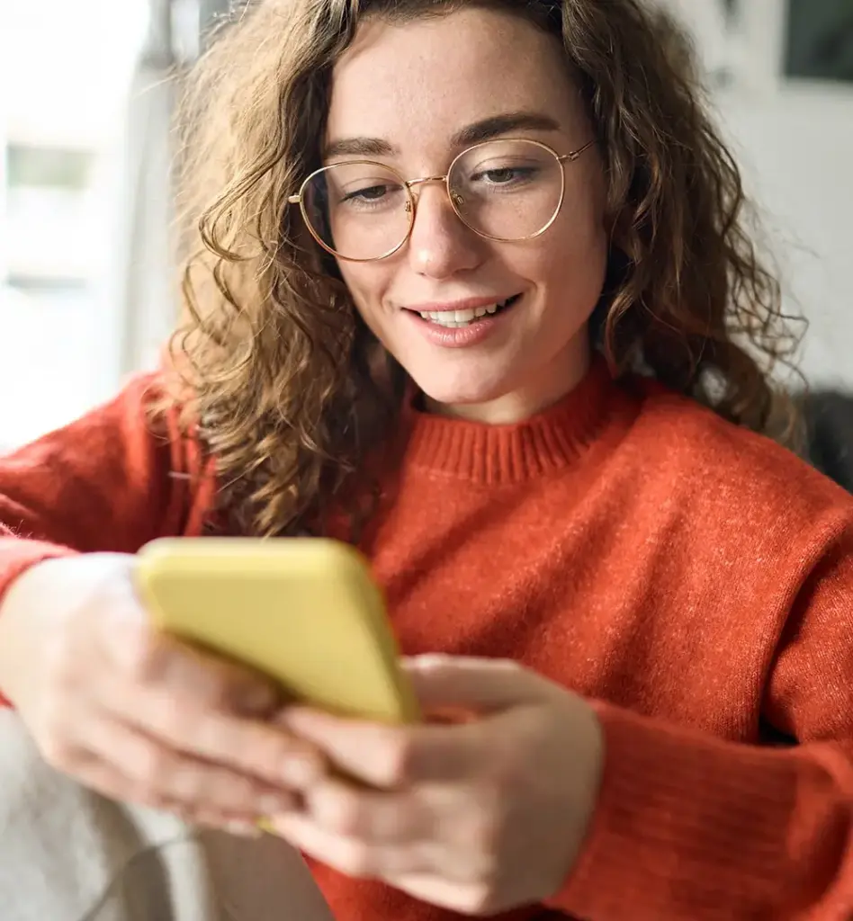 Das Bild zeigt eine junge Frau in einem rotorangen Pullover. Sie trägt eine Brille und blick auf ihr Smartphone. Das Bild wird im Bereich "Krankenkasse? Kannst du jetzt auch per Service-App" verwendet.