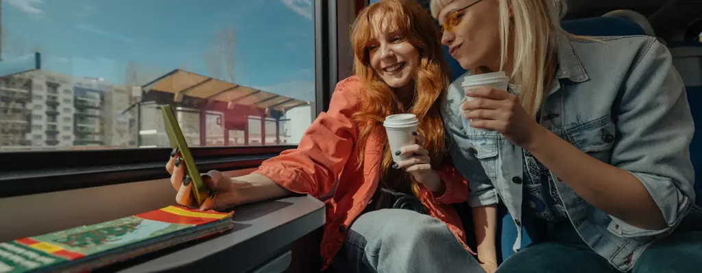 Das Bild zeigt zwei junge Erwachsene, die in einer Bahn sitzen. Sie haben jeweils einen Kaffeebecher in der Hand und machen ein Selfie. Das Bild wird im Bereich "Reiseschutzimpfung für dein nächstes Abenteuer" verwendet.