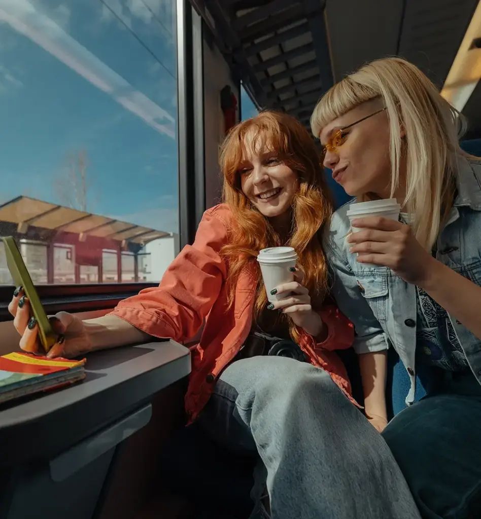 Das Bild zeigt zwei junge Erwachsene, die in einer Bahn sitzen. Sie haben jeweils einen Kaffeebecher in der Hand und machen ein Selfie. Das Bild wird im Bereich "Reiseschutzimpfung für dein nächstes Abenteuer" verwendet.