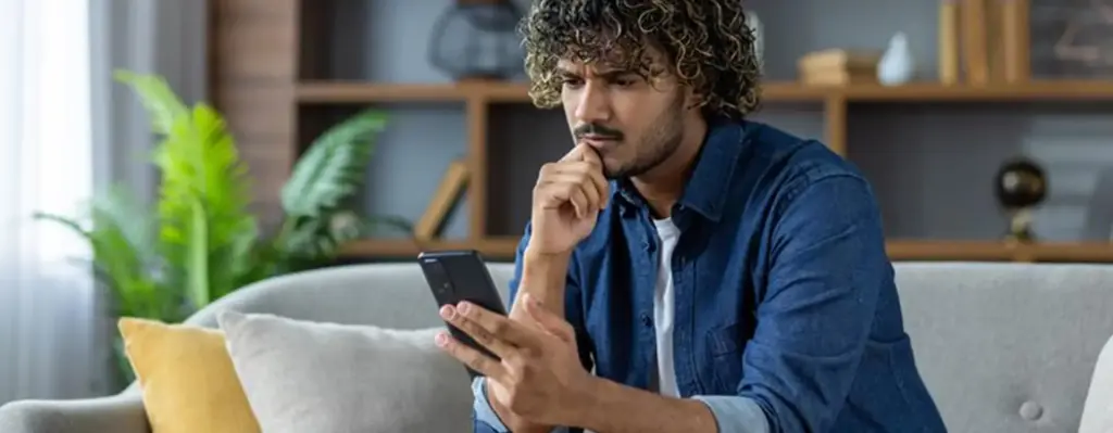 Ein junger Mann sitzt nachdenkend auf einem Sofa und blickt auf sein Smartphone.