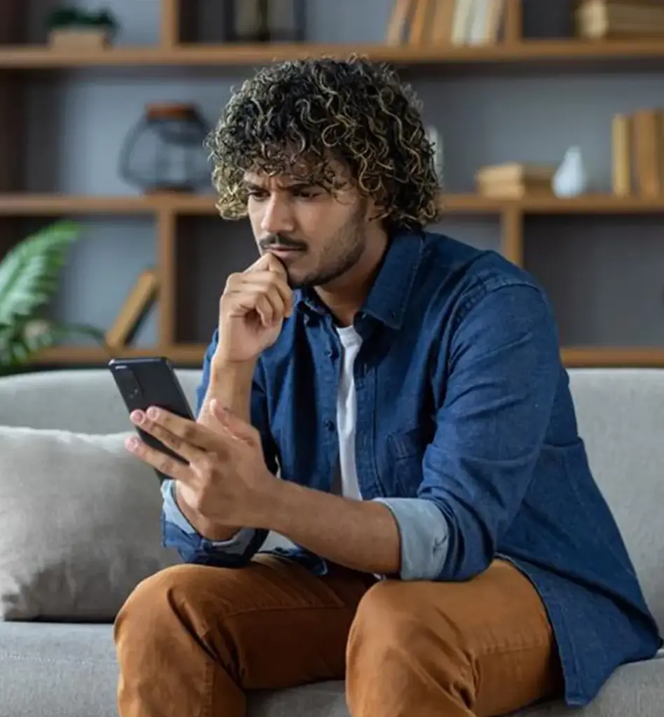 Ein junger Mann sitzt nachdenkend auf einem Sofa und blickt auf sein Smartphone.