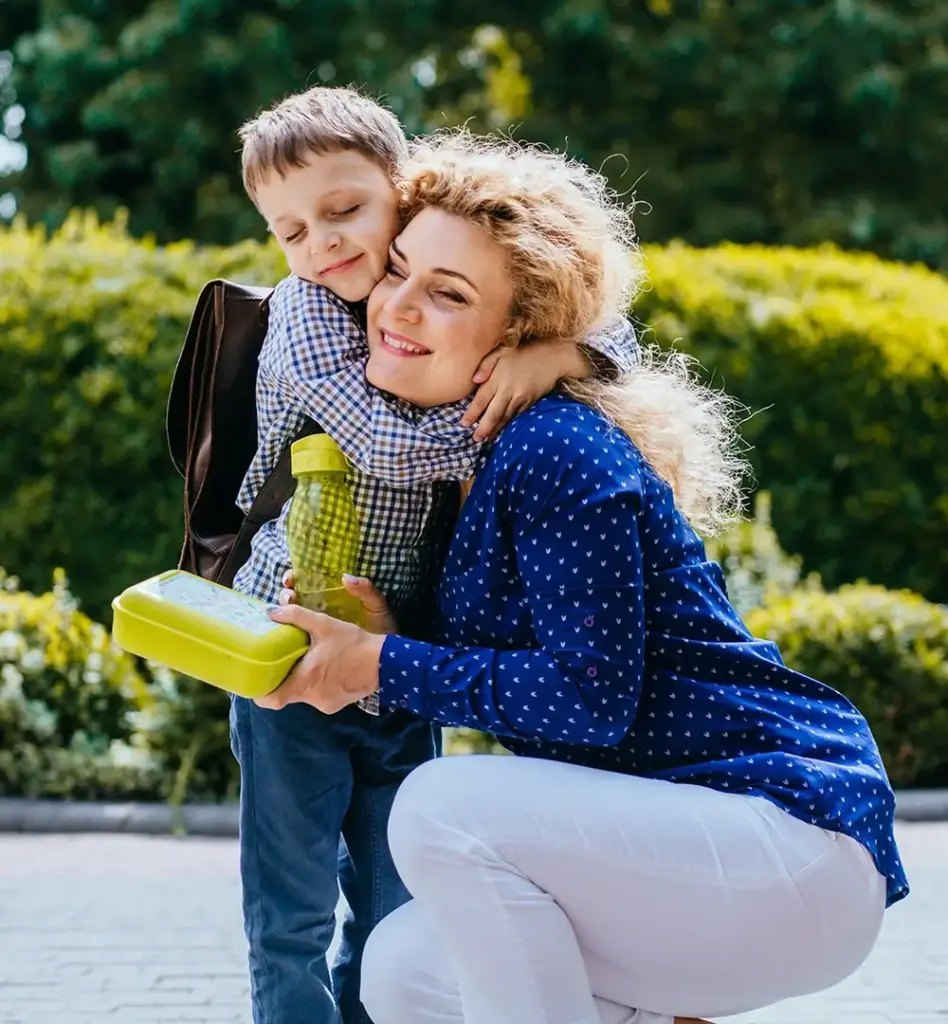 Eine Frau kniet und umarmt ihr Kind. Das Kind hat einen Schulrucksack auf dem Rücken. Die Frau hält eine Flasche und Brotdose fest.