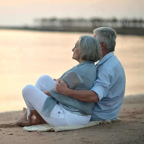 Das Bild zeigt ein älteres Paar, das am Strand auf einer Decke sitzt und aufs Meer blickt. Das Bild wird als Profilbild für Best Ager verwendet.