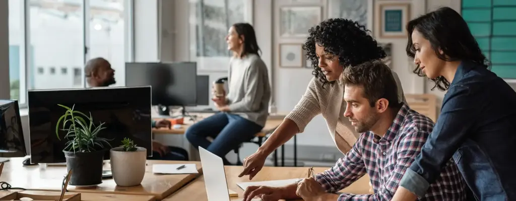 Ein Mann und zwei Frauen schauen gemeinsam in einem Büro auf einen Laptop. Der Mann sitzt, beide Frauen stehen rechts und links von ihm. Eine der beiden Frauen zeigt auf den Bildschirm. Im Hintergund unterhält sich eine Frau mit Kaffeebecher mit einem Mann. Das Bild wird im Bereich "Arbeitnehmer" verwendet.