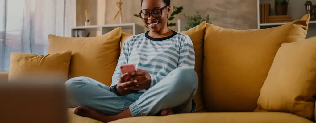Das Bild zeigt eine Frau die im Schneidersitz auf einem gelben Sofa sitzt und auf ihr Handy blickt. Sie trägt eine Brille und lacht. Das Bild wird im Bereich "Digitale Angebote" verwendet.