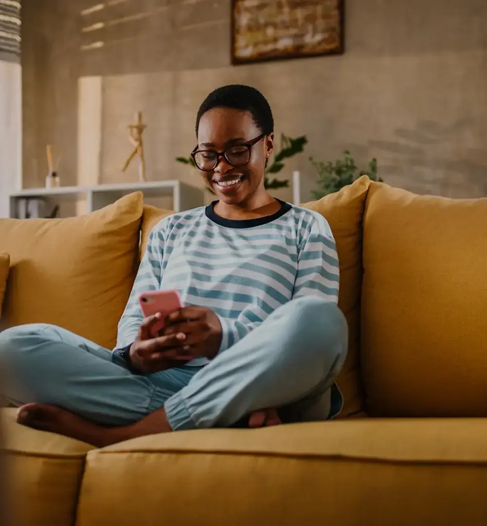 Das Bild zeigt eine Frau die im Schneidersitz auf einem gelben Sofa sitzt und auf ihr Handy blickt. Sie trägt eine Brille und lacht. Das Bild wird im Bereich "Digitale Angebote" verwendet.