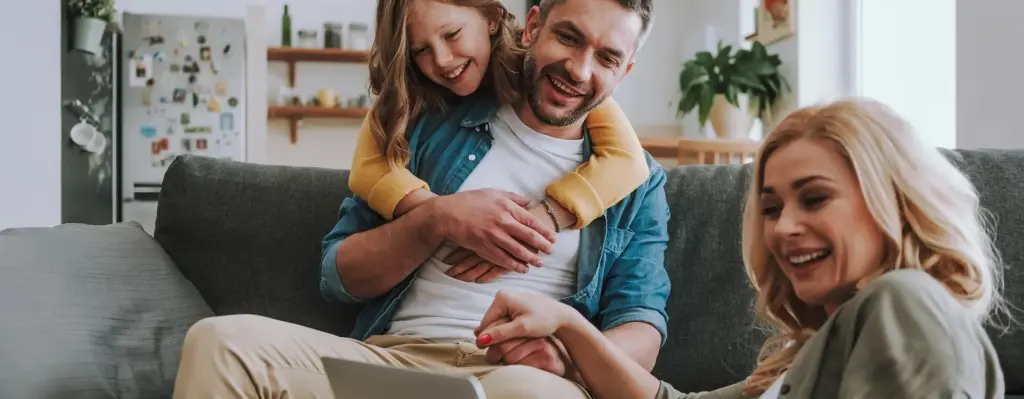 Das Bild zeigt einen Mann, der auf einer Couch sitzt. Im Vordergrund sitzt noch eine Frau mit einem Laptop. Der Mann und die Frau halten Händchen. Ein Mädchen umarmt den Mann von hinten. Alle lachen. Das Bild wird im Bereich "Leistungsübersicht" verwendet.
