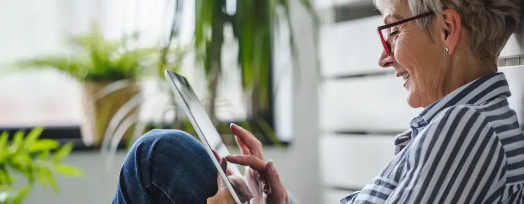 Das Bild zeigt eine Frau, die lacht. Sie trägt eine Brille und eine gestreifte Bluse. Sie sitzt und schaut auf ein Tablet. Das Bild wird im Bereich "Mitgliedermagazin Digital" verwendet.