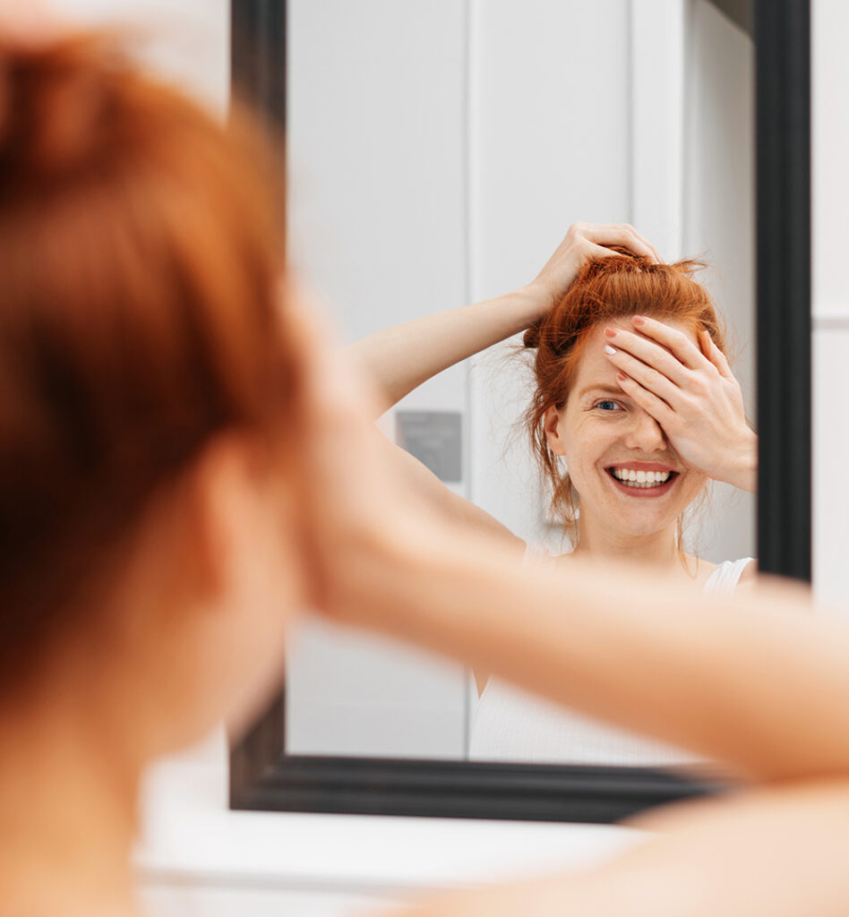 Eine Frau steht vor einem Spiegel und hält sich lachend ein Auge zu. Die andere Hand liegt auf ihrem Kopf. Das Bild wird im Magazin "Perfekt? Lieber nicht" verwendet.
