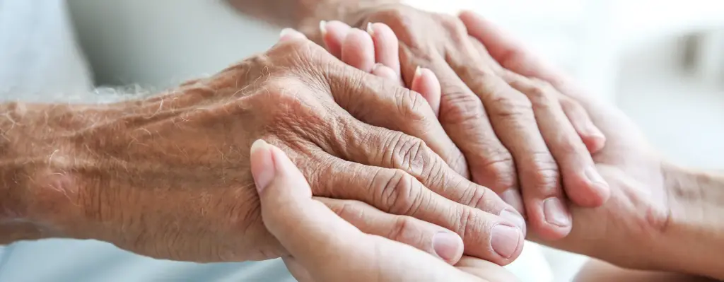 Das Bild zeigt Hände in der Nahaufnahme. Eine jüngere Person hält die Hände einer älteren Person schützend fest. Das Bild wird in dem Bereich "Pflegeversicherung" verwendet.