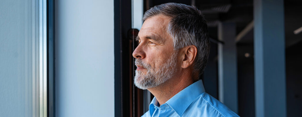 Ein älterer Mann steht an einem Fenster und blickt in die Ferne. Das Bild wird im Magazin "Scharf sehen, klar denken" verwendet.