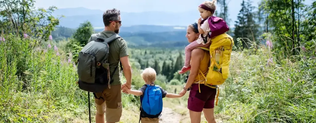 Eine vierköpfige bestehend aus Vater, Mutter, Sohn und Tochter mit Wanderausrüstung steht auf einem Wanderweg. Der Sohn hält die Hande der Eltern, die Tochter sitzt auf den Schultern der Mutter. Beide Eltern schauen sich an. Das Bild wird im Bereich "Schutzimpfung" verwendet.