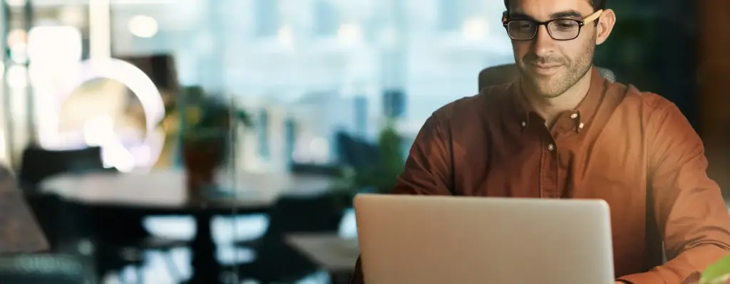 Das Bild zeigt einen Mann mit Brille und Hemd an einem Schreibtisch. Er arbeitet an einem Laptop. Das Bild wird im Bereich "Selbstständige" verwendet.