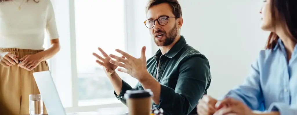 Das Bild zeigt einen gestikulierenden Mann, der mit weiteren Menschen an einem Tisch sitzt. Es spiegelt eine Arbeitssituation wieder. Das Bild wird im Bereich "Die Top-Services der energie-BKK für Arbeitgeber" verwendet.