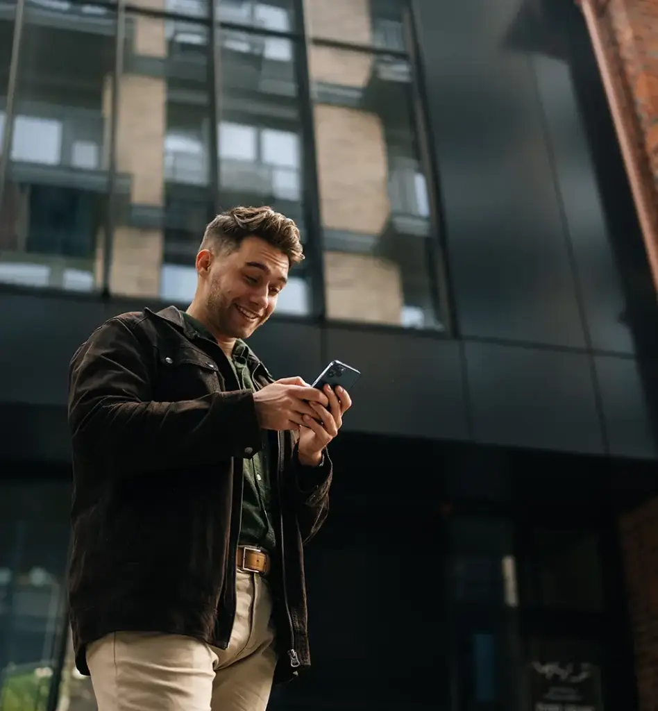 Das Bild zeigt einen jüngeren Mann, der vor einem Gebäude steht. Er blickt auf sein Handy. Die Kameraperspektive ist aufschauend auf den Mann. Das Bild wird im Bereich "Über uns" verwendet.