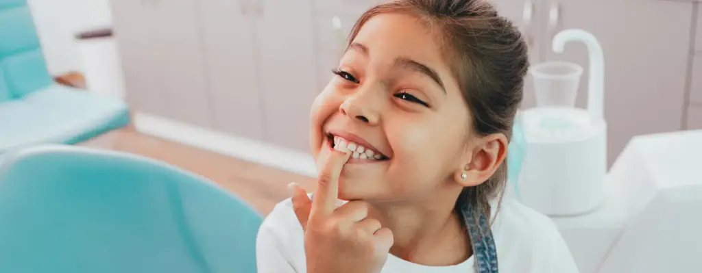 Ein Mädchen sitzt seitlich auf einem Behandlungsstuhl beim Zahnarzt. Sie zeigt mit ihrem Zeigefinger auf ihre Zähne. Dabei lacht sie mit zugekniffenen Zähnen. Das Bild wird im Magazin "Zahngesundheit - von klein auf" verwendet.