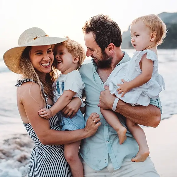 Man sieht eine vierköpfige Familie bestehend aus einer Frau, einem kleinen Jungen, einem Mann und einem kleinen Mädchen. Die Eltern stehen am Strand nebeneinander und lachen. Sie haben jeweils ein Kind auf dem Arm. Das Bild wird als Profilbild für Familien verwendet.