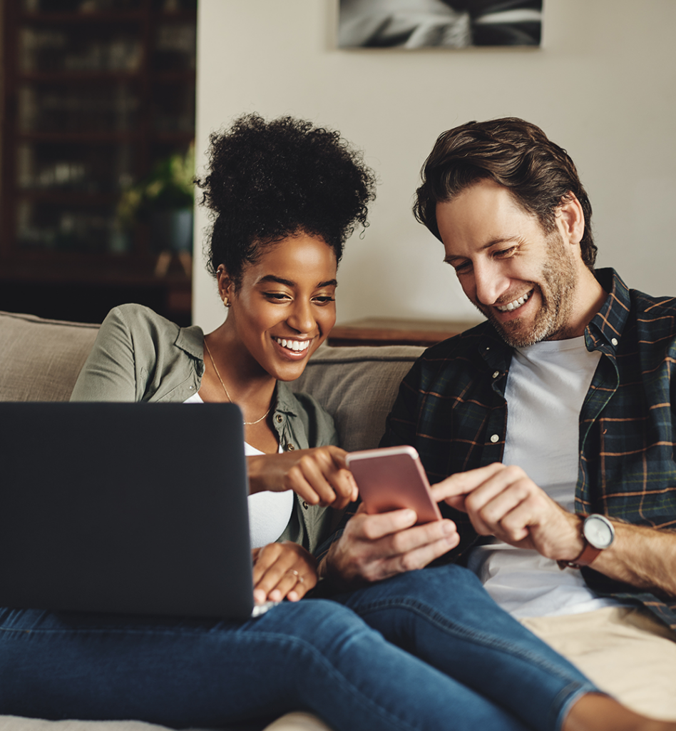 Zu sehen ist ein junges Paar welche zusammen in ein Laptop und in ein Smartphone schauen