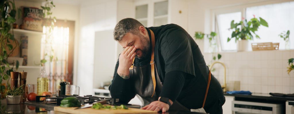 Zu sehen ist ein Mann der erschöpft in der Küche steht