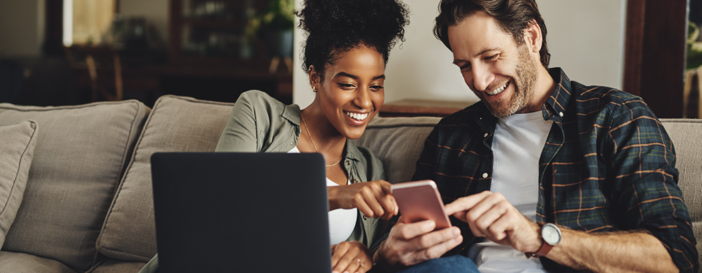 Zu sehen ist ein junges Paar welche zusammen in ein Laptop und in ein Smartphone schauen