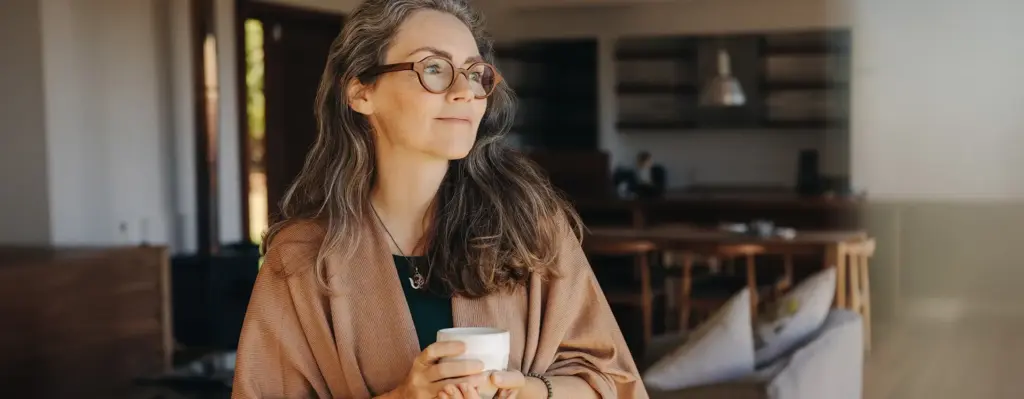 Eine Frau schaut verträumt nach draußen, in der Hand hält sich eine Tasse warmen Tee