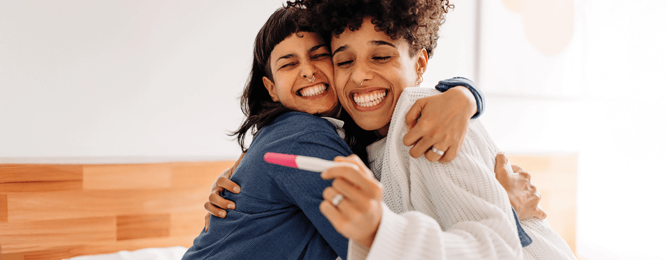 Das Bild zeigt ein Paar bestehend aus zwei Frauen, die sich über einen positiven Schwangerschaftstest freuen