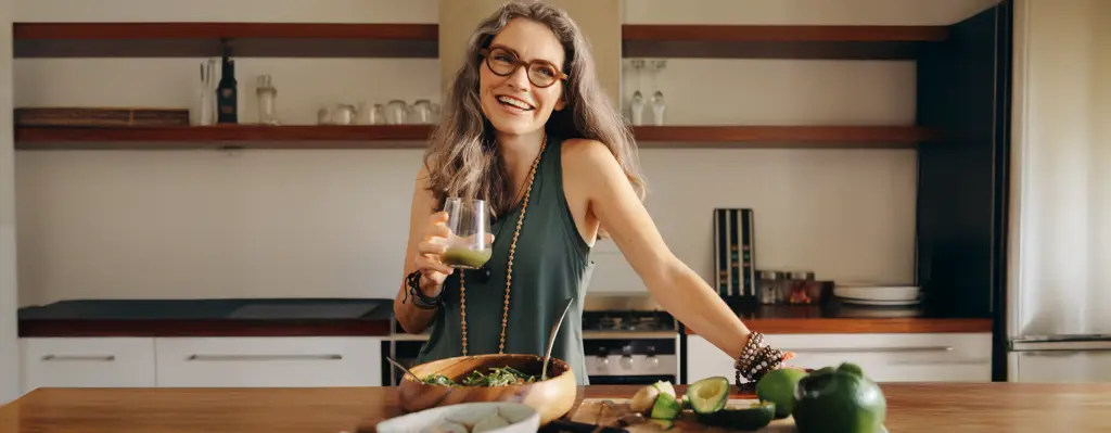 Eine Frau mittleren alters steh lächelnd in der Küche mit einem Glas Saft in der Hand