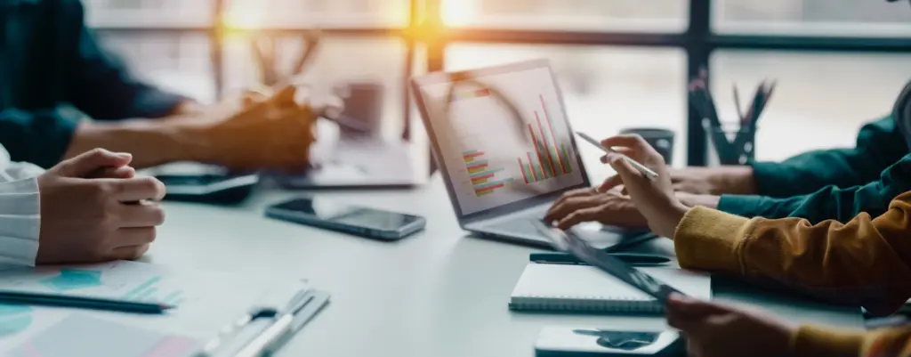 Man sieht einen aufgeklappten Laptop auf einem Schreibtisch in einer Besprechung