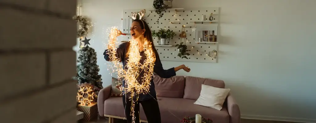 Frau mit Rentiergeweih-Haarreif tanzt ausgelassen im weihnachtlich dekorierten Wohnzimmer – in eine leuchtende Lichterkette gewickelt, singt sie fröhlich.