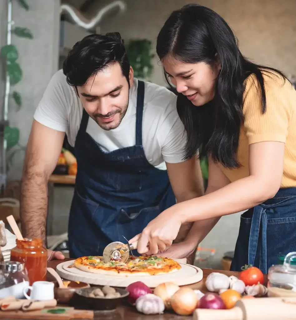 Eine Frau und ein Mann stehen an einem Tisch und die Frau schneidet eine Pizza mit einem Pizzaschneider.