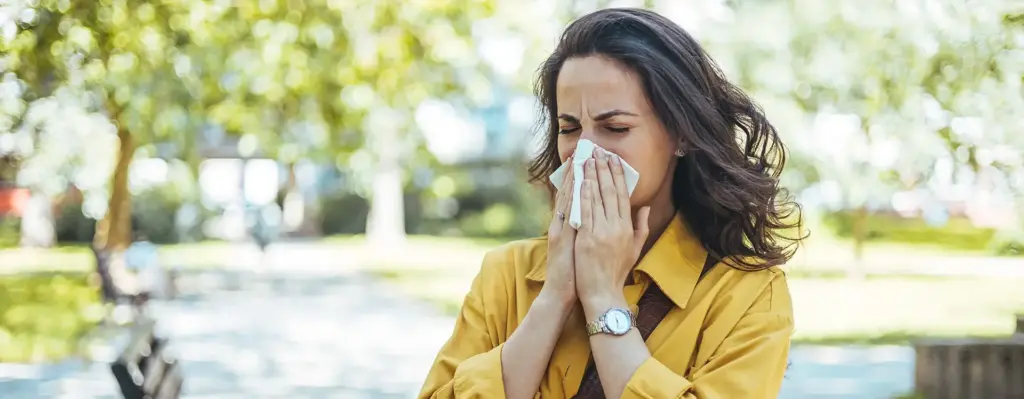 Eine Frau in gelber Jacke putzt ihre Nase mit einem Taschentuch. Sie ist draußen in einem Park.