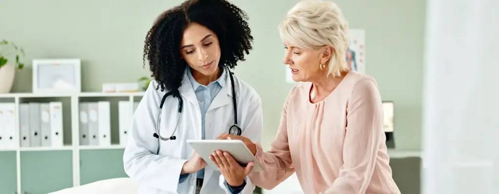 Eine Frau sitzt auf einer Untersuchungsliege. Neben ihr steht eine Ärztin mit einem Tablet und zeigt der Patientin etwas.