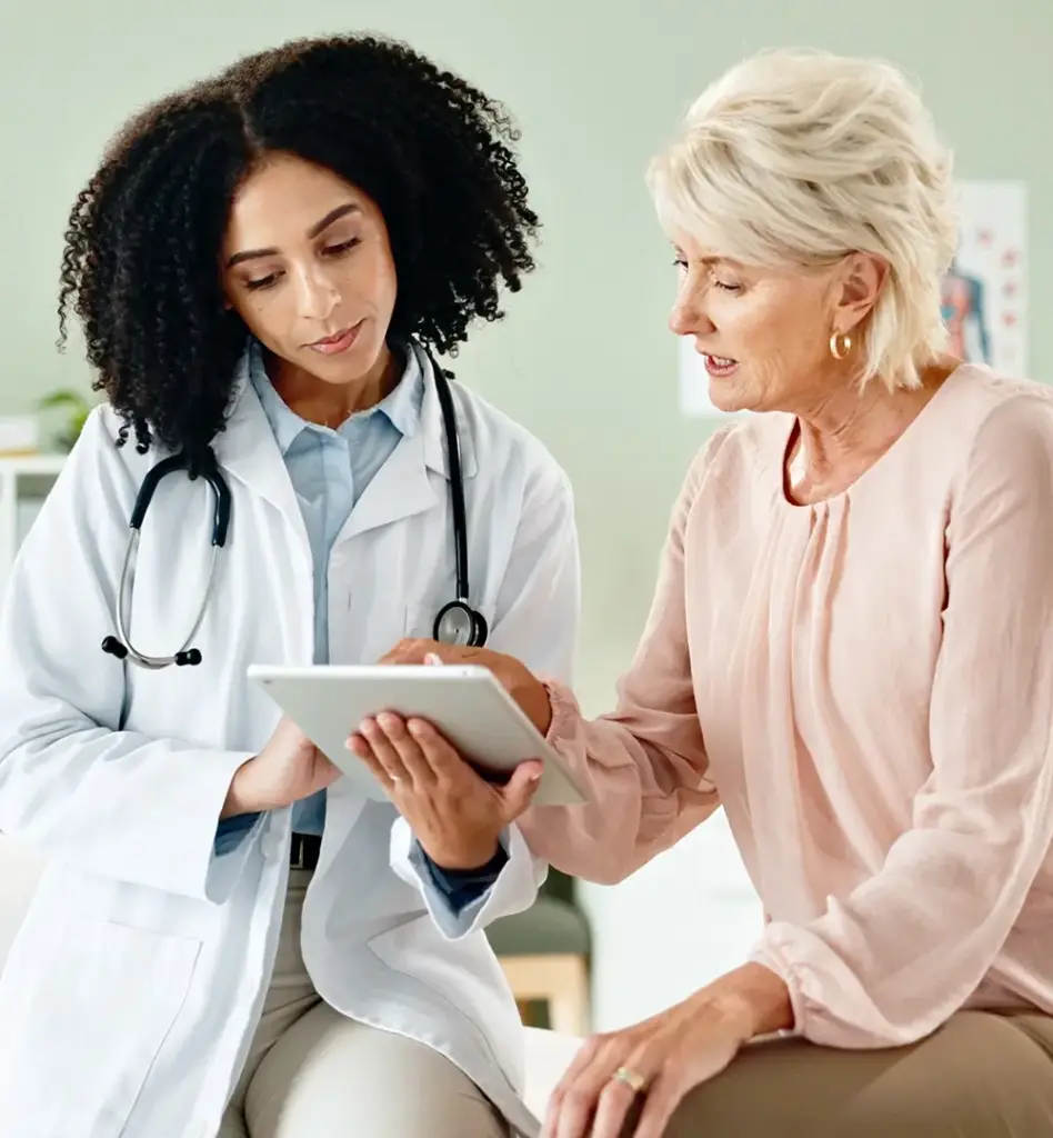 Eine Frau sitzt auf einer Untersuchungsliege. Neben ihr steht eine Ärztin mit einem Tablet und zeigt der Patientin etwas.