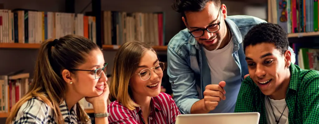 Das Bild zeigt eine Gruppe von Studenten die in einer Bibliothek vor einem Laptop sitzen