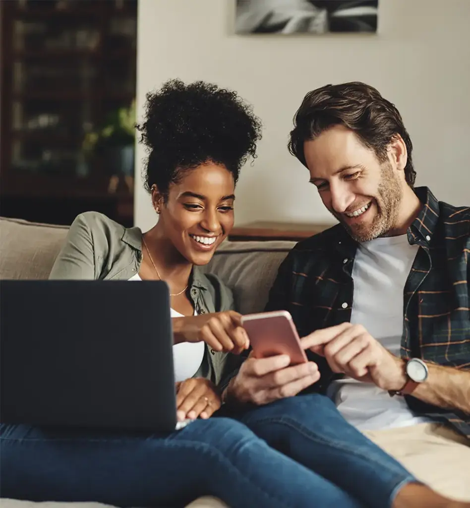 Zu sehen ist ein junges Paar welche zusammen in ein Laptop und in ein Smartphone schauen