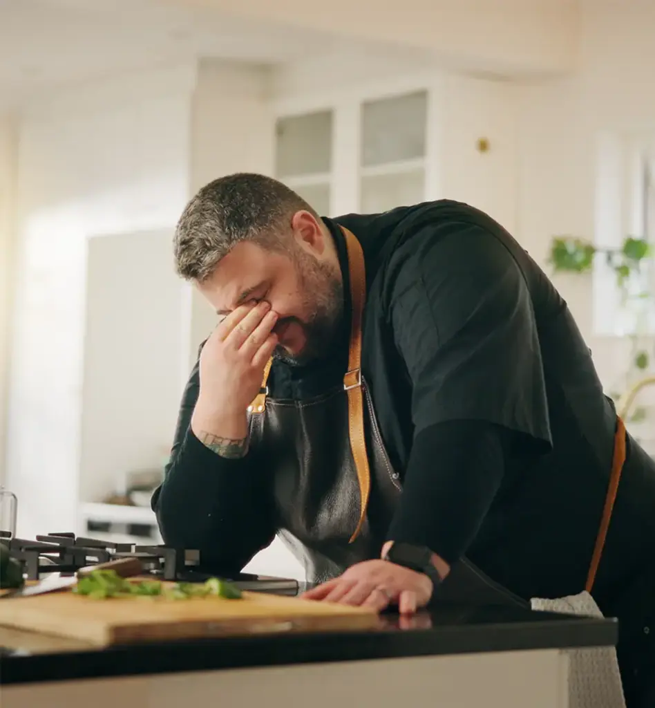 Zu sehen ist ein Mann der erschöpft in der Küche steht