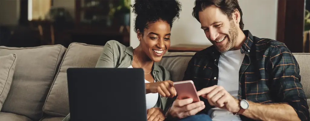 Zu sehen ist ein junges Paar welche zusammen in ein Laptop und in ein Smartphone schauen