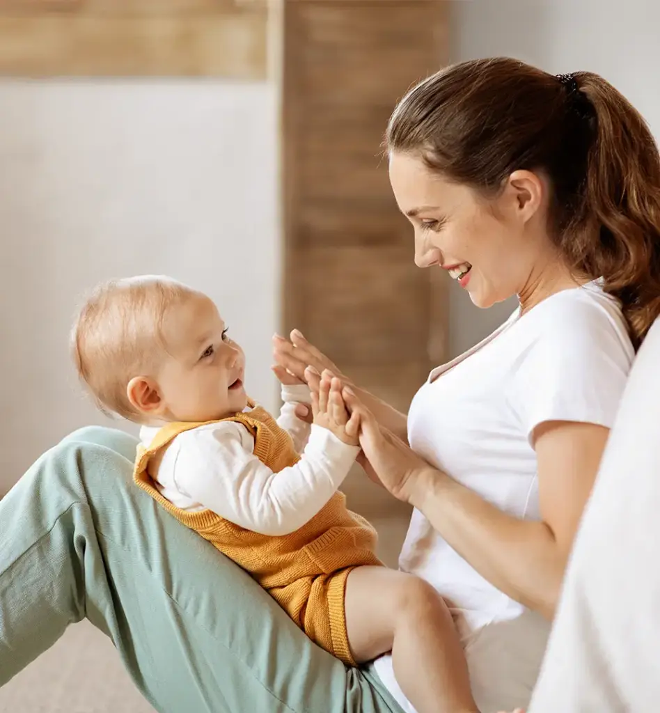 Eine Mutter sitzt auf dem Boden, Beine angewinkelt auf Ihrem Schoss sitzt ihr kleines Baby