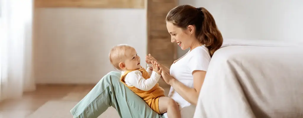 Eine Mutter sitzt auf dem Boden, Beine angewinkelt auf Ihrem Schoss sitzt ihr kleines Baby