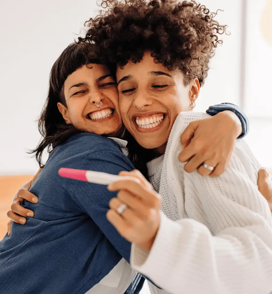 Das Bild zeigt ein Paar bestehend aus zwei Frauen, die sich über einen positiven Schwangerschaftstest freuen
