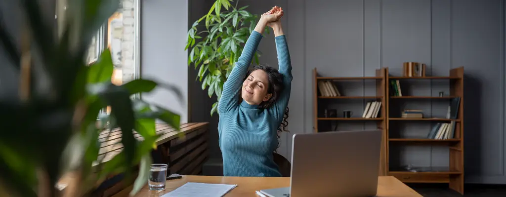 Eine Frau sitzt im Homeoffice an ihrem Laptop und streckt die Arm nach oben