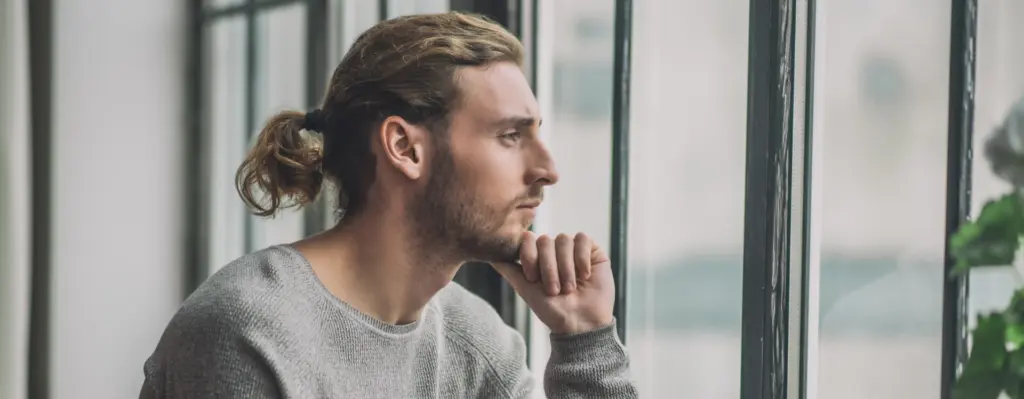 Ein junger Mann sitze nachdenklich vor dem Fenster, seinen Kopf in die Hand gestützt
