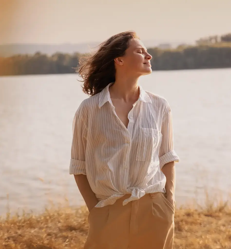 Eine Frau steht an einem See und genießt mit geschlossenen Augen die Seeluft